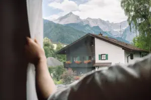 Uitzicht vanuit de salon Een hand houdt een gordijn voor een raam met bergen op de achtergrond.