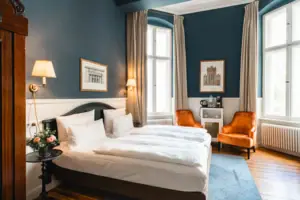 Stijlvolle hotelkamer in het HENRI Berlin met donkerblauwe muren, hoge plafonds, twee oranje fauteuils en een elegant tweepersoonsbed in een historische ambiance.