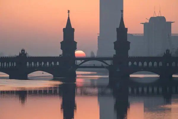 Silhouet van de Oberbaumbrücke in Berlijn bij zonsopgang, met de torens en de roodachtige zon weerspiegeld in het kalme water van de Spree.