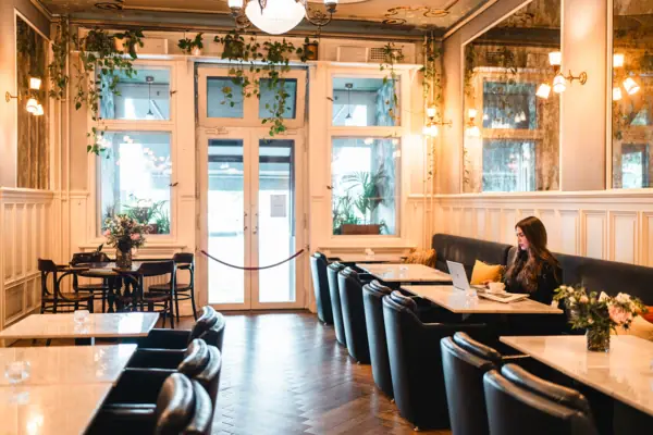Een jonge vrouw werkt op haar laptop in de stijlvolle hotellobby van het HENRI Berlin, omringd door klassieke houten lambrisering, marmeren tafels en hangende groene planten in de Belle Époque stijl.