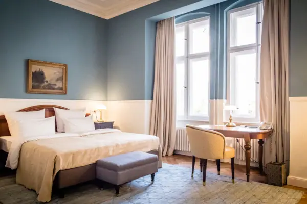 Stijlvolle hotelkamer in het HENRI Berlin met lichtblauwe muren, een tweepersoonsbed, een antiek houten bureau en hoge ramen in de klassieke stijl van een oud gebouw.