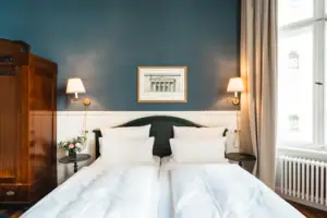 Vooraanzicht van een stijlvol tweepersoonsbed in de HENRI Berlijn met diepblauwe muur, klassieke houten kledingkast, symmetrische wandlampen en een ingelijste architectuurprint.