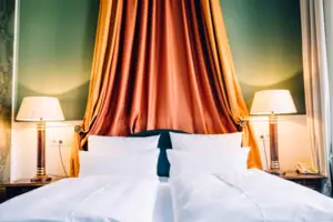 HENRI Hotel Berlin Kamerdetails: baldakijn Detailfoto van een statig hotelbed in het HENRI Berlin met een prachtige oranje-gouden baldakijn tegen een groene muur en klassieke nachtlampjes.