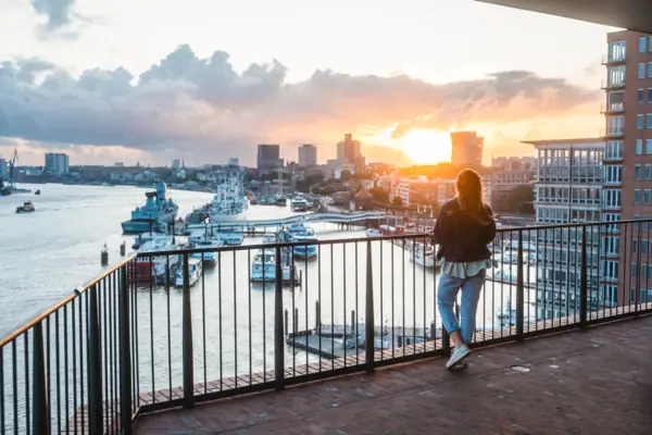 Hamburg Elphie Een vrouw staat op een balkon en kijkt uit over het water met boten op de achtergrond.