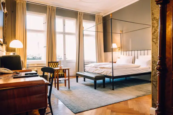 Ruime, lichte hotelkamer in het HENRI Berlin met een klassiek hemelbed, visgraat parketvloer en stijlvolle vintage meubels voor hoge ramen.