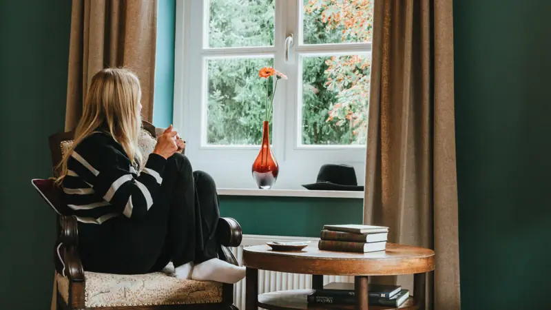 Kamers in het HENRI Kitzbühel Een vrouw zit op een stoel en kijkt uit het raam.